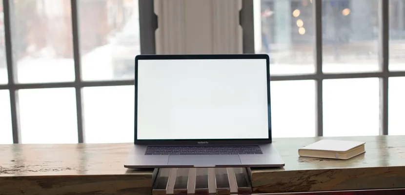 A laptop with a blank white screen open on a wooden desk beside a closed notebook, in front of a tall window